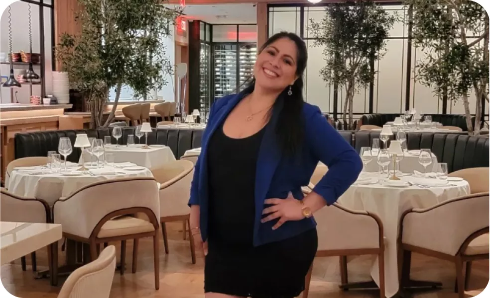 Chef Magaly Ponce standing confidently in a restaurant, wearing a blue blazer and black dress, smiling at the camera with a well-set dining area in the background.