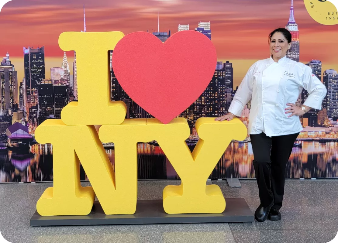Chef Magaly Ponce standing next to a large 'I ❤️ NY' sign in New York, smiling and wearing her chef's coat.