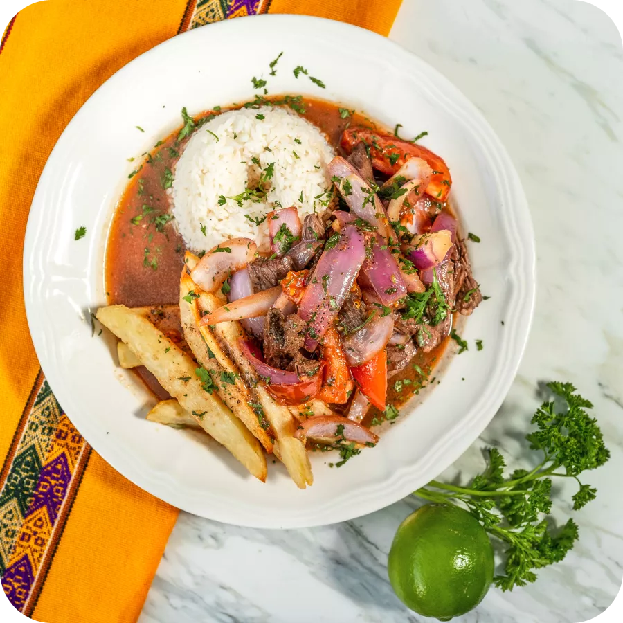 Plate of Lomo Saltado with beef, onions, tomatoes, served with rice, fries, and garnished with cilantro and lime.